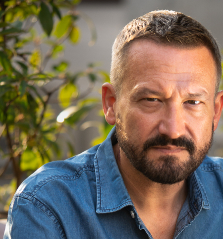 Portrait of Franck Finance-Madureira. A person with short dark blonde hair and a beard is wearing a denim shirt and sitting at a table in a garden setting.