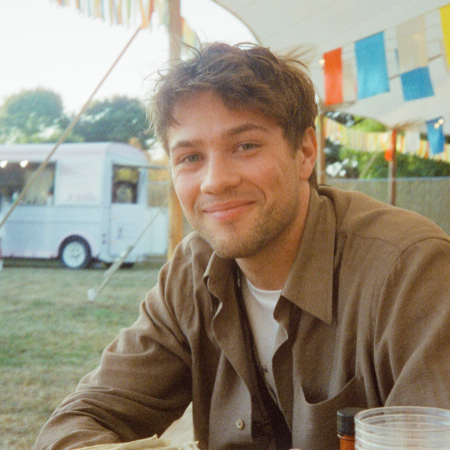 Portrait of Connor Jessup. A person with a dark-blonde 3-day beard and a stylish crew cut sits on a bench at a festival.